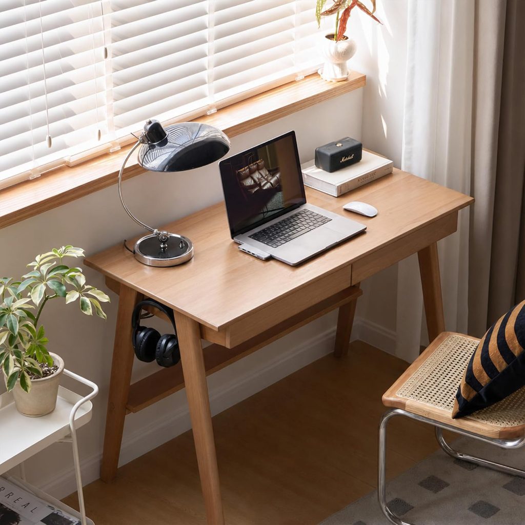 Solid wood desk positioned by a window with laptop, lamp, and headphones hanging from the built-in hook